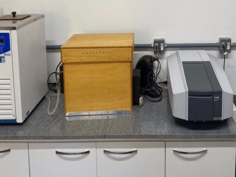 Fotocatalise_A Descrição da imagem. A imagem apresenta uma fotografia de uma bancada de um laboratório com três equipamentos em cima. O primeiro da esquerda para a direita é denominado banho térmico, equipamento retangular de cor branca, com detalhes do visor em azul, apenas uma parte dele está visível na foto. No meio, uma caixa em madeira denominada de caixa de degradação. E a direita o espectrofotômetro, equipamento também retangular de cores branca e cinza, mais baixo que os outros dois. Abaixo da bancada é possível observar 5 portas de um armário branco, todas fechadas, com puxadores prateados. Ao fundo parede branca, onde tomadas e fios estão visíveis. Fim da descrição